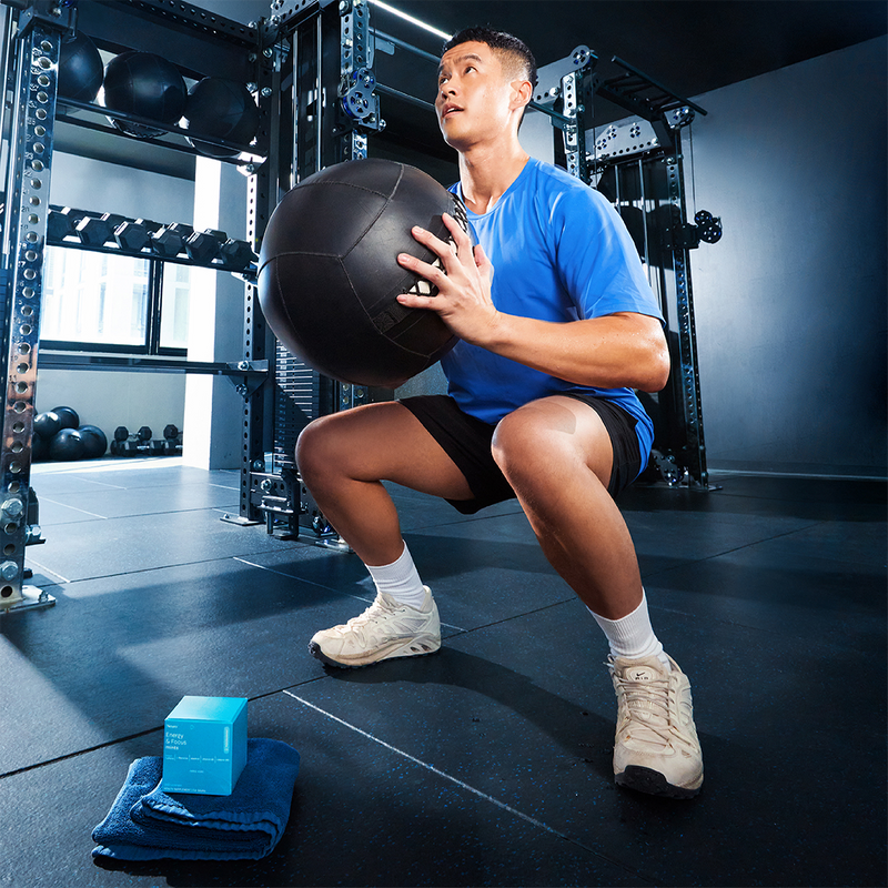 Man squatting with a medicine ball in a gym with Neuro Energy & Focus Mints pack on the floor